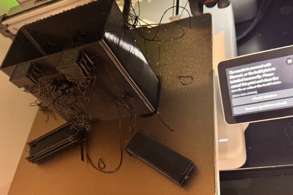 A badly failed black print tangled across the A1 build plate while the touchscreen displays a nozzle-filament warning